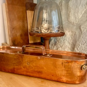 May include: An elongated, oval-shaped copper fish poacher with a matching lid. The vessel features two brass side handles and a central brass handle on the lid, all secured with rivets. The polished copper surface reflects light across its metallic body.