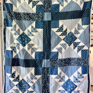 May include: A rectangular quilt featuring a patchwork design in various shades of blue and cream. The pattern consists of geometric blocks, including triangles and squares, with diverse fabric prints such as polka dots, plaid, and floral motifs.