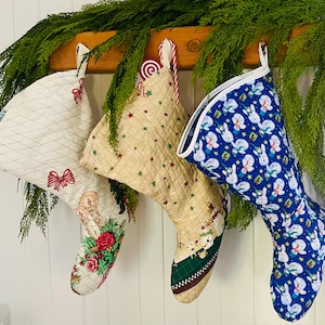May include: Three Christmas stockings hanging on a wooden shelf. The stocking on the left is white with a red bow and a floral pattern. The middle stocking is beige with red stars and a snowman pattern. The stocking on the right is blue with a white snowman pattern.