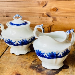 May include: A white porcelain sugar bowl with a lid and a creamer. Both pieces have blue floral designs and accents. The sugar bowl has a handle on the lid. The creamer has a curved spout and a handle.