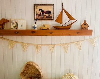 Peut inclure: Une étagère en bois avec un fond blanc, décorée d'un voilier, d'une voiture jouet, d'une image de chevaux, d'une petite maison en bois et de deux livres de Peter Rabbit. Une guirlande en dentelle blanche est suspendue sous l'étagère.