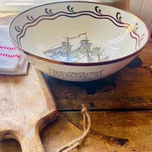 Peut inclure: Un bol en céramique blanc avec un bord marron et une illustration détaillée d'un voilier. Le bol a une bordure décorative marron et dorée autour du bord et une citation en bas.