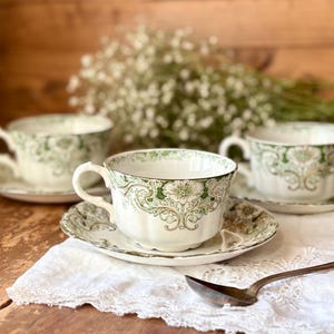May include: Three antique teacups and saucers with a green floral design and gold trim. The teacups are white with a scalloped edge and a delicate handle. The saucers match the teacups, with a similar floral pattern. A silver spoon rests on a white cloth.