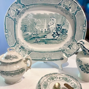 May include: A vintage tea set comprising a serving platter, sugar bowl, small plate, and a creamer. The teal and white set features a detailed landscape scene on the platter. The sugar bowl and creamer have matching teal trim. A spoon and knife rest on the small plate.