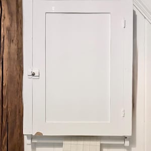 May include: A rectangular white wall-mounted cabinet with a single door and a small metal latch. A white towel with a green embroidered leaf pattern and a central letter C hangs from a horizontal bar attached to the bottom of the cabinet frame.