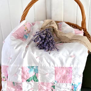 May include: A wicker basket with a curved handle contains a white quilt with pink floral patchwork. A bunch of dried lavender tied with a tan ribbon sits on the quilt. The quilt features pink, white, and blue floral patterns.