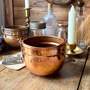 May include: A display of antique copper pots and glass jars on a wooden surface. A tall white candle is in a brass holder, and a small black and white photograph rests on the table. The scene has a rustic, vintage aesthetic.