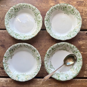 May include: Four white ceramic bowls with a green and gold floral design around the rim. One bowl has a silver spoon resting inside. The bowls are arranged on a wooden surface.