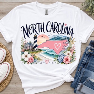 Peut inclure: Un t-shirt blanc à manches courtes avec une impression graphique portant le texte NORTH CAROLINA en bleu foncé. Le motif comprend un phare, une carte rose de la Caroline du Nord avec un cœur blanc, une plage, des montagnes et des fleurs.