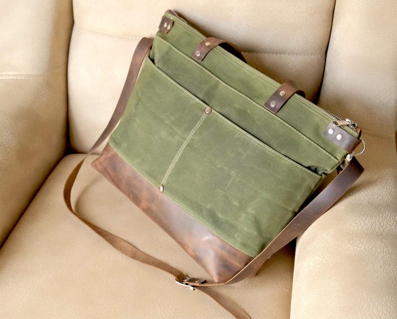 May include: A green and brown canvas and leather tote bag with a long shoulder strap. The bag has multiple pockets and a zipper closure.