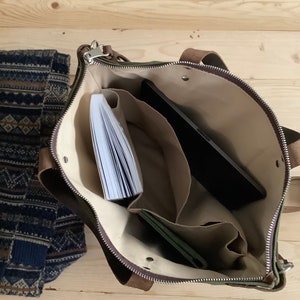 May include: A green canvas tote bag with brown leather accents and a zipper closure. The bag is open and has several compartments inside, including a large pocket for a laptop or tablet. There is a book and a wallet inside the bag.