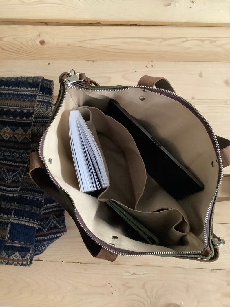 May include: A green canvas tote bag with brown leather accents and a zipper closure. The bag is open and has several compartments inside, including a large pocket for a laptop or tablet. There is a book and a wallet inside the bag.