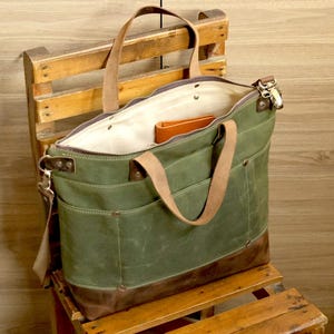 May include: A green canvas tote bag with brown leather accents and a zipper closure. The bag has a large main compartment and a smaller pocket on the front. The bag is sitting on a wooden chair.