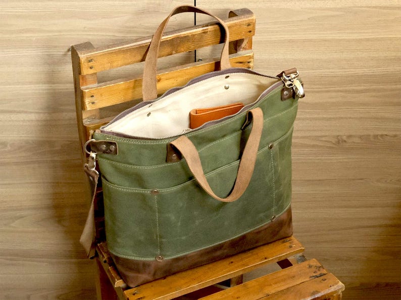 May include: A green canvas tote bag with brown leather accents and a zipper closure. The bag has a large main compartment and a smaller pocket on the front. The bag is sitting on a wooden chair.
