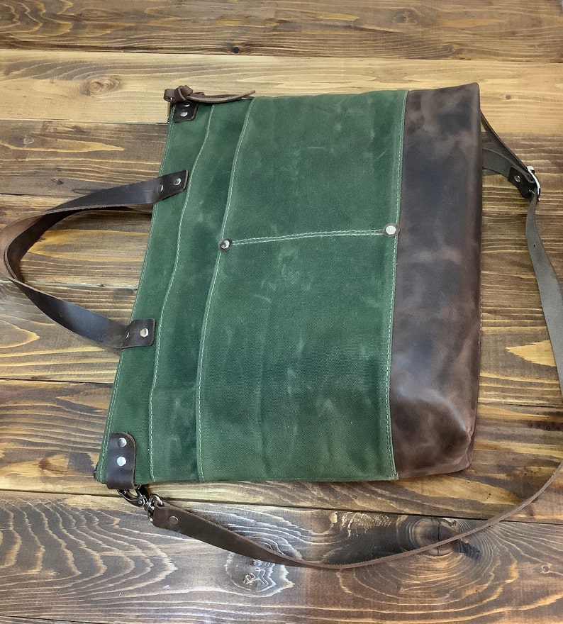 May include: A green canvas tote bag with brown leather accents and a long brown leather strap. The bag has a zipper closure and multiple pockets.