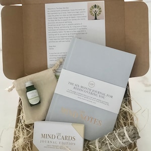 以下が含まれることがあります： セルフケアアイテムでいっぱいの茶色の段ボール箱。箱の中には、ジャーナル、カードセット、エッセンシャルオイルのボトル、セージのスマッジスティック、小さな袋が入っています。ジャーナルのタイトルは「The Six-Month Journal for Rediscovering You」。カードのタイトルは「Mind Cards Journal Edition」。エッセンシャルオイルのボトルには「Breathe Deep White」とラベルが貼られています。
