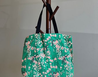 May include: A green fabric tote bag featuring a repeating pattern of black monkeys and pink cherry blossom flowers. The bag has two black fabric handles attached to the top edge, allowing it to be carried or hung from a hook for storage.