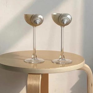 May include: Two clear glass candle holders with a seashell design. The candle holders are on a light brown wooden table.