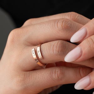 May include: A rose gold ring engraved with the words "LIFE" and "LOVE". The ring wraps around the finger, featuring a minimalist design. The image shows the ring on a hand with manicured nails. The ring is a stylish piece of jewellery.