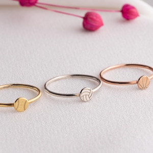 May include: Three delicate rings in gold, silver, and rose gold, each featuring a sports-themed charm. The charms depict a baseball, a volleyball, and a basketball. The rings are displayed on a white surface with pink floral accents.
