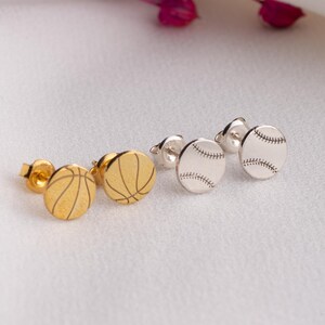 May include: Two pairs of stud earrings, one pair in gold featuring a basketball design and one pair in silver featuring a baseball design. Each earring is circular with engraved lines representing the texture of the respective sports balls.