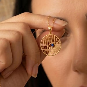 May include: A gold-colored pendant necklace featuring a circular, openwork design with a grid pattern. A small, round, blue gemstone is set in the center of the pendant. The necklace is held up by a hand.