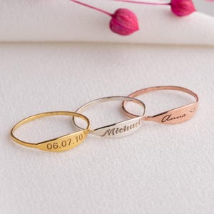 Könnte beinhalten: Drei zarte Ringe in Gold, Silber und Roségold. Jeder Ring hat eine personalisierte Inschrift: "06.07.10", "Michael" und "Anna". Die Ringe werden auf einer weißen Oberfläche mit rosa floralen Akzenten präsentiert.