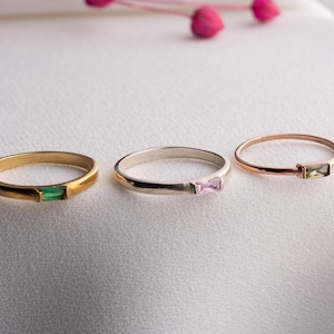 以下が含まれることがあります： 長方形の宝石が付いた3つの繊細なリング。リングは金、銀、ローズゴールドで、それぞれ異なる色の石（緑、ピンク、黄緑）が付いています。白い表面にピンクの花のアクセントを添えて展示されています。