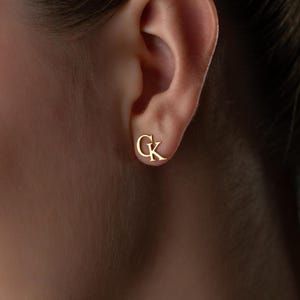 May include: A close-up of a gold-colored stud earring with the letters "G" and "K" in a script font. The earring is positioned on an ear, with the focus on the jewelry. The background is blurred.