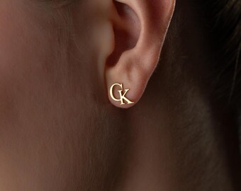 May include: A gold-coloured stud earring featuring the letters "G" and "K" in a script font. The earring is set against an ear, with the focus on the jewellery. The background is blurred.