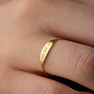 May include: A gold ring with the engraved date "06.07.10" is shown on a finger. The ring has a smooth, polished surface and a simple, elegant design. The ring is a warm, yellow colour.