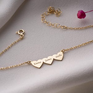 Op de afbeelding: Een goudkleurige armband met drie hartvormige bedels. Elk hart is gegraveerd met een naam: Emma, James en Stella. De armband ligt op een witte, getextureerde ondergrond, met een kleine roze bloem op de achtergrond.