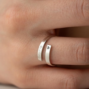 May include: A silver wrap ring, engraved with the name "Sophia" and a small heart symbol. The ring's design is minimalist, with the name and heart adding a personal touch. The ring is worn on a finger.