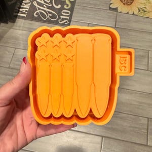 May include: An orange silicone mold featuring a raised design of an American flag where the stripes are replaced by vertical bullet shapes and the canton contains six stars. The letters ISC are embossed on a small rectangular tab on the right side.