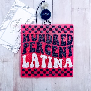 May include: A pink wooden sign with black and white checkered border. The sign says "HUNDRED PERCENT LATINA" in black and white glitter letters.