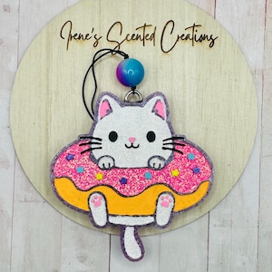 May include: A cat-shaped air freshener with a donut design. The white cat has pink ears and paws, black whiskers, and is surrounded by a pink glitter donut with yellow frosting. The text "Irene's Scented Creations" is visible on the wooden background.