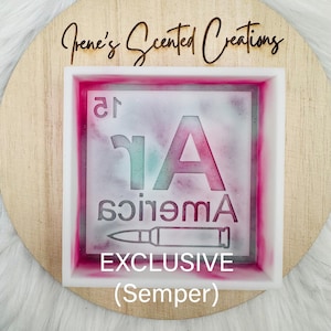 May include: A square resin mold with the word "America" in a stencil design, a bullet, and the word "EXCLUSIVE" is displayed. The mold is set on a wooden circle with the text "Irene's Scented Creations". The mold has pink and teal accents.