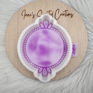 Op de afbeelding: Een witte en paarse mal met een decoratieve rand en een centraal cirkelvormig ontwerp. De mal ligt op een houten oppervlak met de tekst "Irene's Crafted Creations". De achtergrond is een wit, pluizig materiaal.