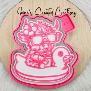 May include: A pink and white layered cookie cutter depicting a Highland cow wearing sunglasses and a flower crown, on a duck-shaped pool float. The cutter has the text "Irene's Scented Creations" on a wooden background.