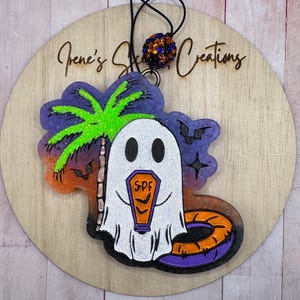 May include: A Halloween-themed air freshener with a white ghost holding a coffin marked "SPF", a palm tree, and a purple and orange sunset. It hangs from a black string with a decorative bead.