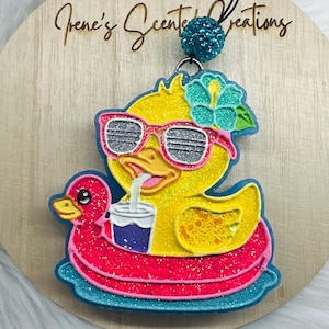 May include: A yellow glitter duck wearing pink sunglasses and a flower, on a pink and blue pool float, drinking from a cup. The text "Irene's Scented Creations" is on the wooden background.