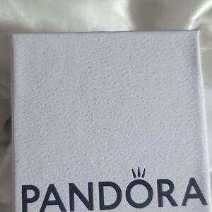 Op de afbeelding: Een vierkant wit sieradendoosje met een textuur op het oppervlak. Het woord PANDORA staat in donkergrijze hoofdletters op de voorzijde gedrukt, met het kenmerkende kroonsymbool van het merk direct boven de letter O in de naam.