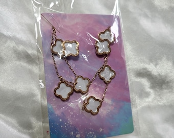 May include: A rose gold-toned chain necklace featuring multiple four-leaf clover charms with white pearlescent centers. The necklace is displayed on a rectangular card with a pink, purple, and blue watercolor design, enclosed in a clear plastic bag.