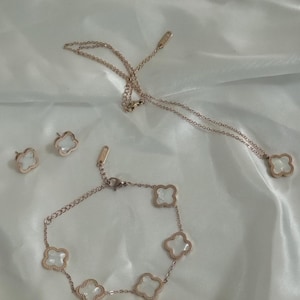 Può includere: Un set di gioielli in oro rosa con un design a trifoglio. Il set comprende una collana, un braccialetto e orecchini. I ciondoli a trifoglio sono realizzati in madreperla bianca.