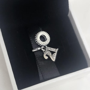 Op de afbeelding: Een zilveren bedel in de vorm van het getal 21, versierd met kleine heldere steentjes. De bedel heeft een ronde bovenring met de inscriptie P2 S925 ALE, wat duidt op sterling zilver, en hangt aan een zwart lint in een doosje.