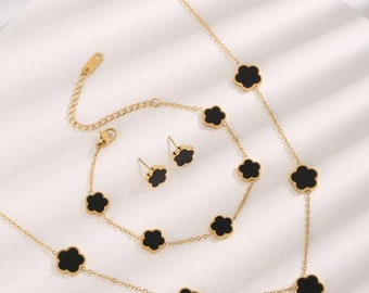 May include: A gold-colored jewelry set featuring a necklace, bracelet, and stud earrings. Each piece is adorned with black clover-shaped accents. The set is displayed on a white surface, showcasing the elegant design.