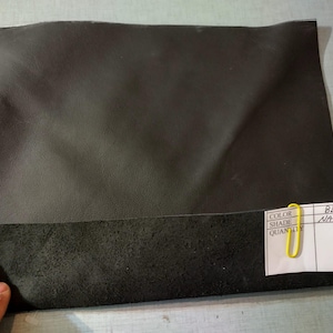 May include: A piece of black leather with a rough, textured bottom edge. A small white tag is attached with a yellow paper clip, with the words "BLACK NATURAL" written on it. The leather is likely for crafting or upholstery.