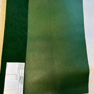 Könnte beinhalten: Ein rechteckiges Stück waldgrünes Leder mit einer strukturierten genarbten Oberfläche und einer glatten Wildlederunterseite. Ein weißes Etikett ist an der unteren linken Ecke befestigt mit dem Text COLOR SHADE GREEN FOREST und QUANTITY 50.