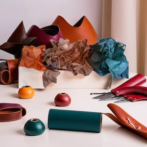 Puede incluir: Una caja blanca llena de recortes de cuero en varios colores, incluyendo marrón, naranja, rojo y azul. También hay tres pequeños recipientes redondos con tapas, uno amarillo, uno rojo y uno verde. Un juego de herramientas de trabajo del cuero se coloca sobre una superficie blanca.