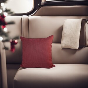 May include: A close-up of a cream-colored couch with a red throw pillow and a white throw blanket. The couch is in a room with a window in the background.